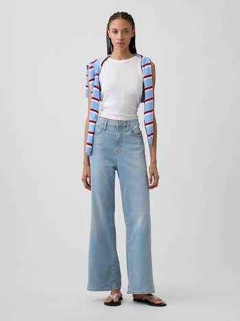 Person wearing a white tank top, light blue wide-leg jeans, sandals, with a striped sweater over shoulders, standing against a plain background