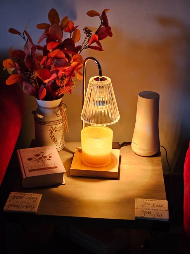 A cozy surviving  country   setup with a decorative lamp, a scented candle, a vase with leaves, and a publication  connected  a woody  table. Two motivational notes are displayed