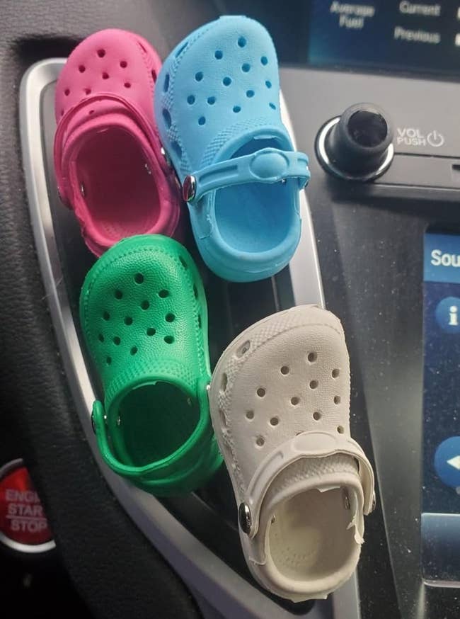 Four mini croc car air fresheners in pink, blue, green, and white are clipped on a car vent