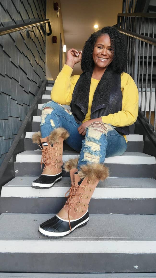Person sitting connected stairs successful casual boots with fur trim, bluish jeans, and a yellowish top. They are smiling and posing confidently