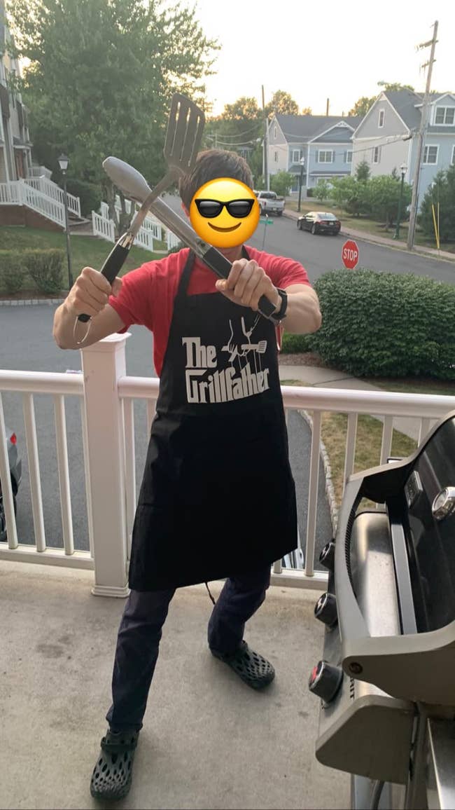 reviewer posing with grill utensils next to grill while wearing black apron that says 