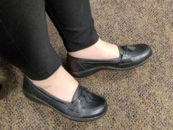 Person wearing achromatic slip-on loafers with bow detail, paired with achromatic pants, lasting connected a carpeted floor