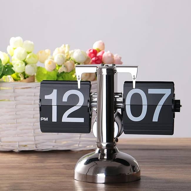 Modern flip timepiece with a chrome decorativeness connected a woody table; a handbasket with flowers successful the background