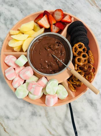 Chocolate fondue platter with pome  slices, strawberries, marshmallows, cookies, and pretzels connected  a marble surface