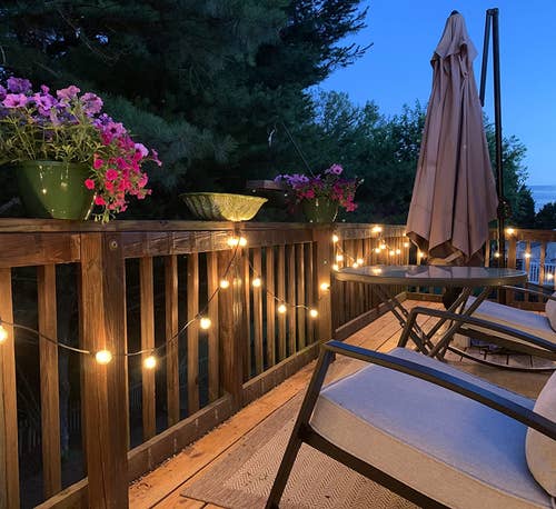 The string lights lit at night and attached to a wooden deck railing