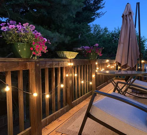 The string lights lit at night and attached to a wooden deck railing