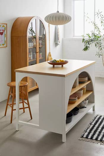 Modern kitchen island with open shelving, displaying dishes and a fruit bowl. Nearby, a wooden cabinet with glass doors and two wooden bar stools