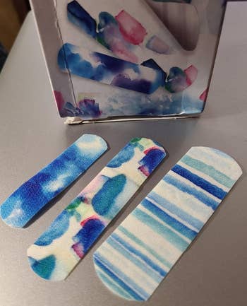 Three patterned adhesive bandages displayed on a surface near a box suggesting artistic, colorful designs for stylish wound care