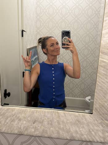 Person successful reflector selfie wearing a sleeveless button-up top. They are posing with manus signs. Bathroom inheritance with patterned wallpaper
