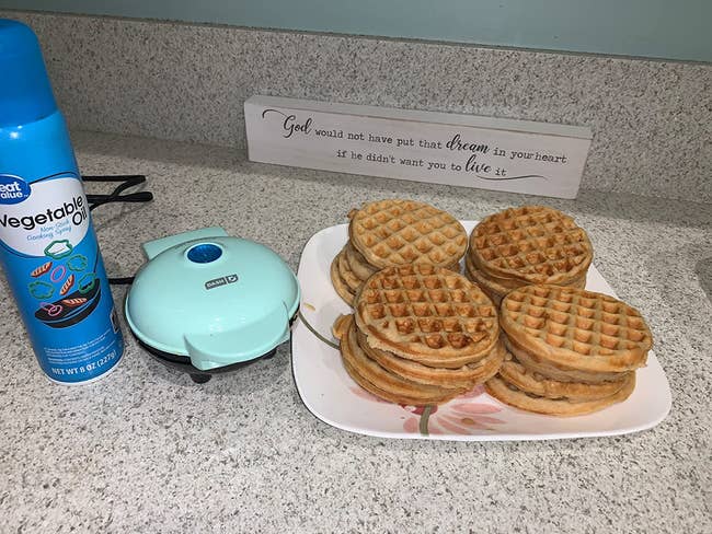 A reviewer's aqua waffle maker next to a plate filled with 16 waffles that have been made