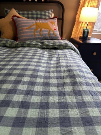 Plaid bedding with a decorative pillow featuring a tiger plan  connected  a neatly made furniture  adjacent  to a lamp-lit nightstand