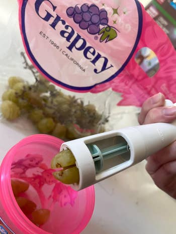 A hand using a kitchen tool to slice grapes above a bowl, with a bag of grapes labeled 