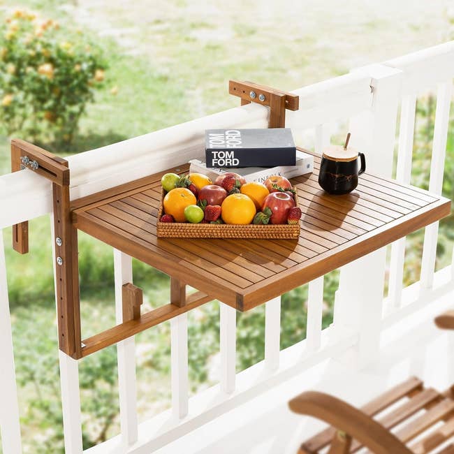 Wooden balcony table with a fruit basket, book, and cup on top, mounted on a white railing