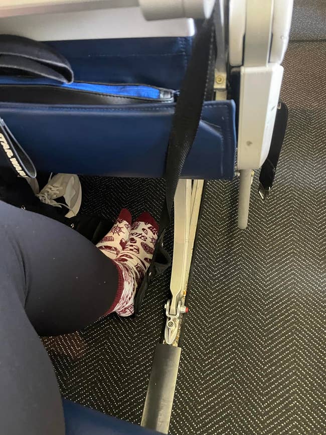 Person wearing black leggings and patterned socks sitting in an airplane seat, showing their feet and part of the tray table mechanism