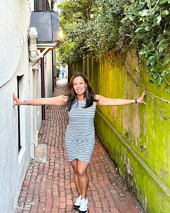 Woman in a striped dress with arms outstretched in a narrow alley, wearing sneakers. She's smiling at the camera