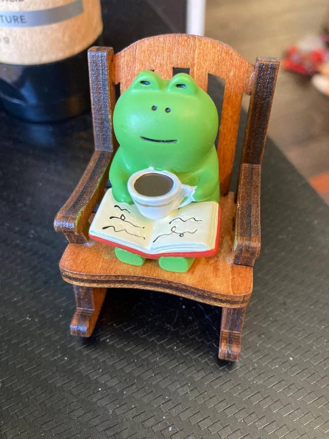 A small frog figurine sits in a miniature wooden chair, holding a book and a coffee cup