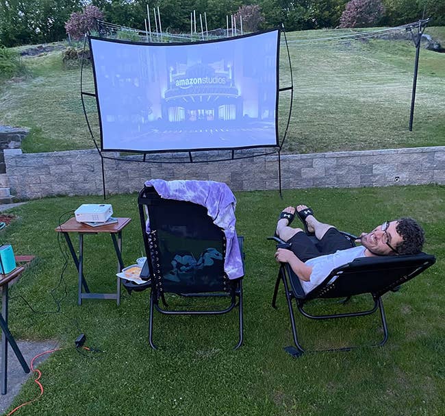 Person relaxing in a chair facing an outdoor movie screen at dusk with a snack table nearby