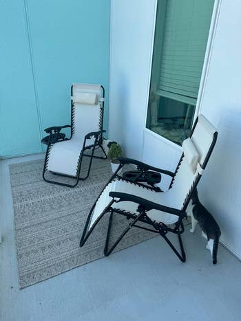 Two patio chairs with an attached table and drink holder, adjacent to a window on an outdoor rug. A cat is partially visible