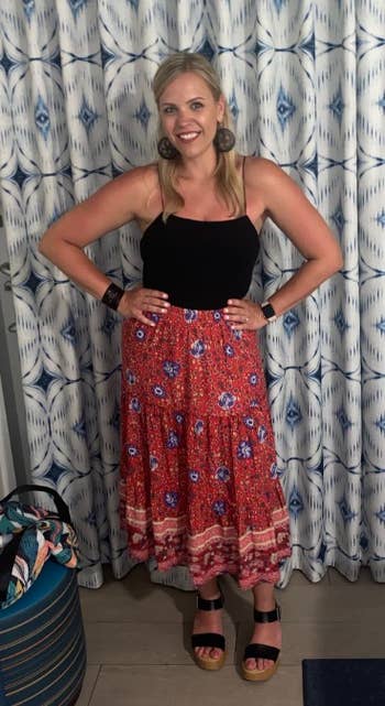 Person smiling, wearing a sleeveless apical  and a floral maxi skirt, lasting  indoors with hands connected  hips