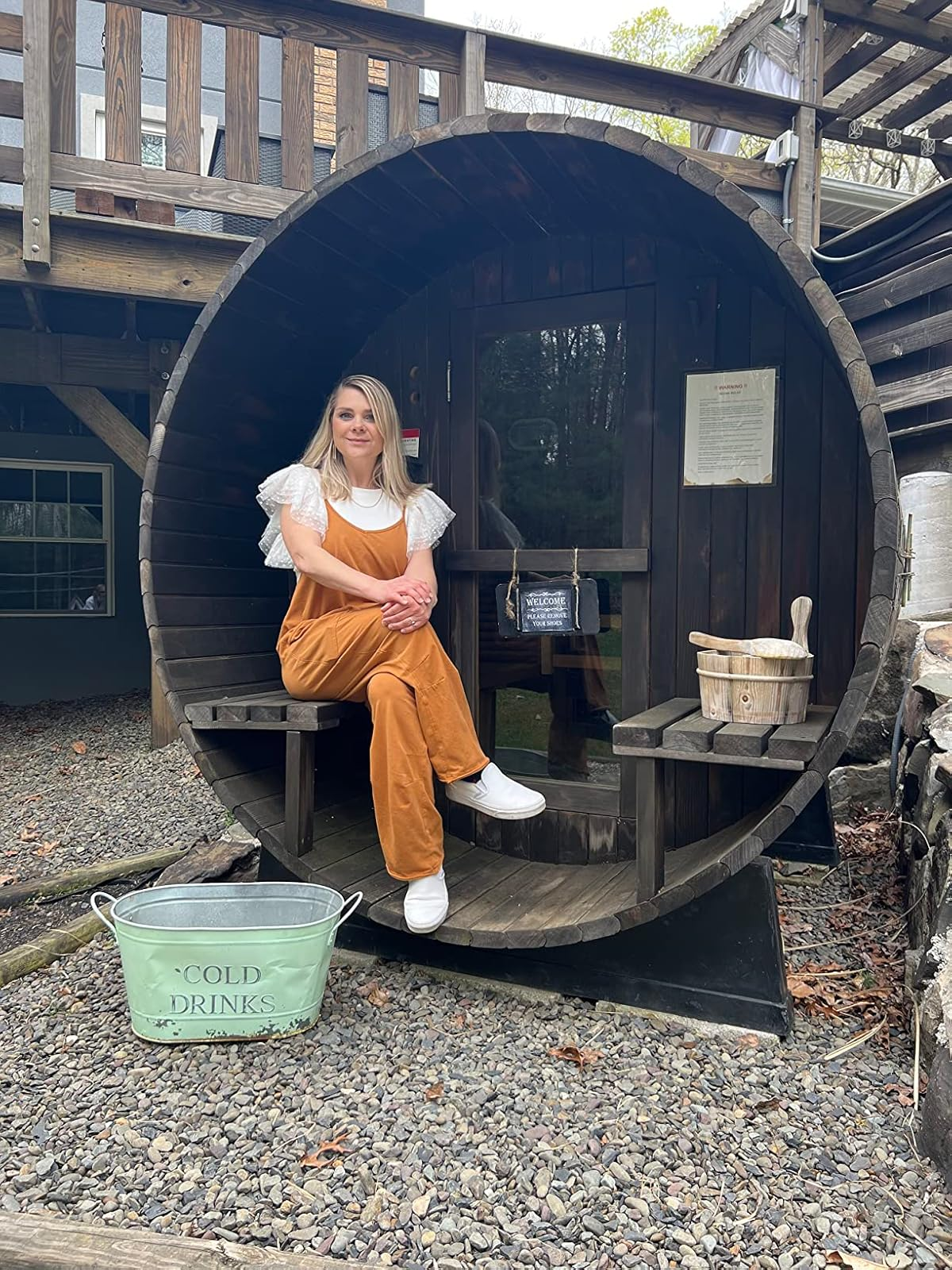 A person sits in a barrel-shaped sauna, wearing casual, relaxed clothing. A bucket labeled 