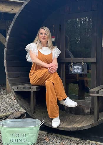 A person sits in a barrel-shaped sauna, wearing casual, relaxed clothing. A bucket labeled