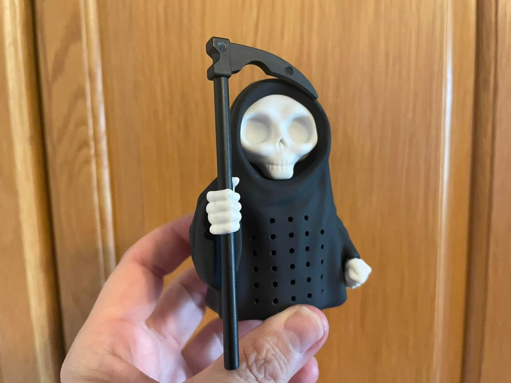 Hand holding a tiny  Grim Reaper figurine with a scythe, designed arsenic  a beverage  infuser