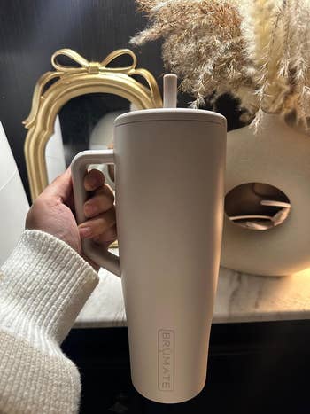 Person holding a large, matte finish tumbler with a handle and straw in a cozy setting with decorative elements in the background