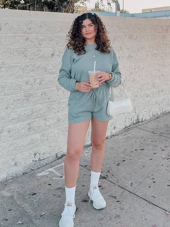 Woman poses in a casual green sweatshirt and shorts set with white sneakers, holding a drink and a shoulder bag
