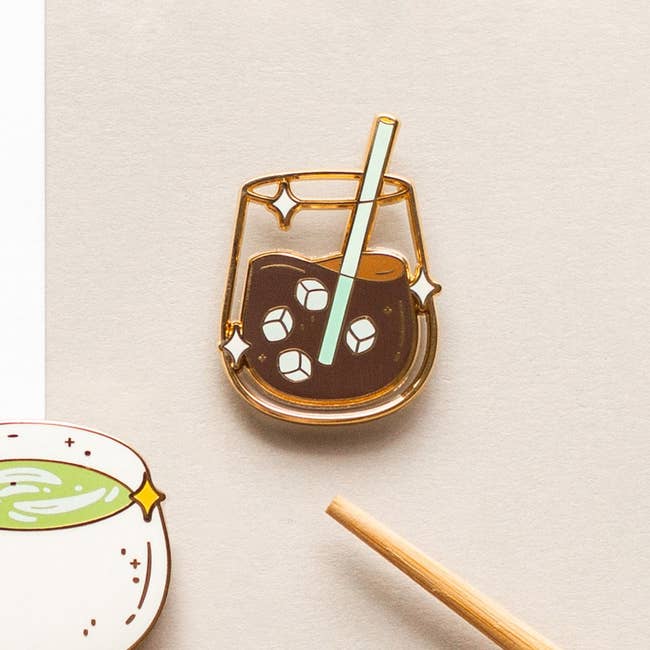 A cute enamel pin depicting an iced java  with a straw and crystal  cubes, styled with a minimalist design