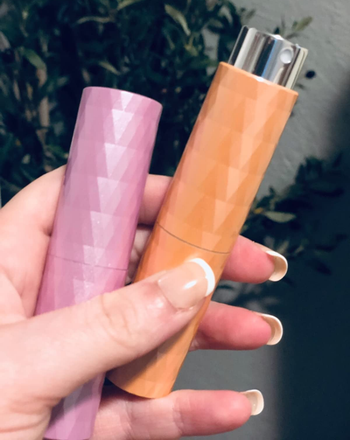 a reviewer holding the shiny pink and orange spray bottles which are roughly the size of a mascara tube