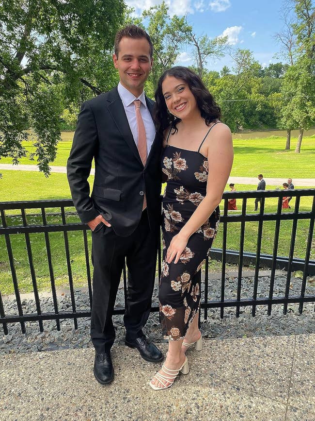 A couple in semi-formal attire: man in black suit and woman in floral dress, posing together outdoors