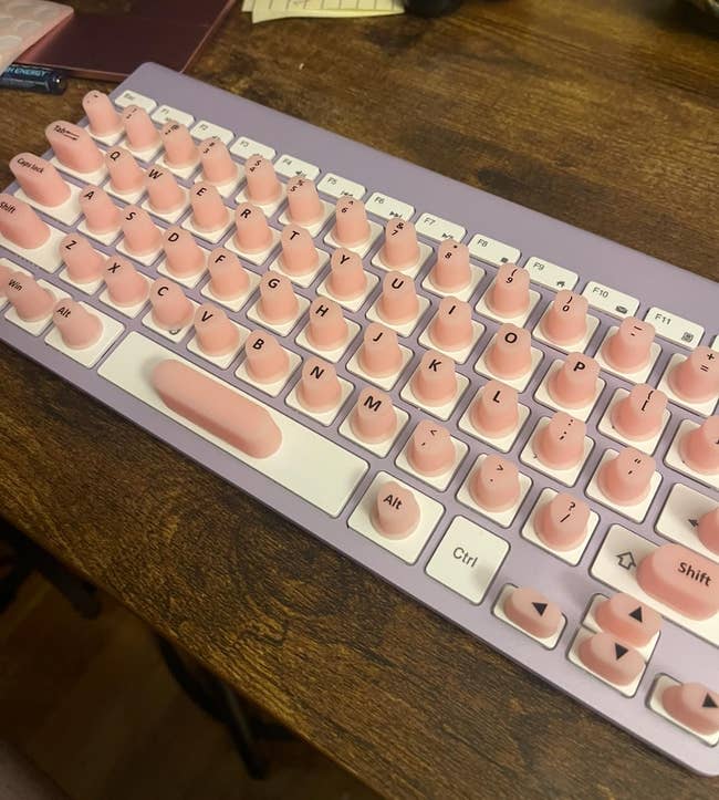 Keyboard mekanis bertema pastel dengan tombol membulat di atas meja kayu, menampilkan aksen merah jambu dan desain penuh gaya. Cocok untuk pecinta belanja