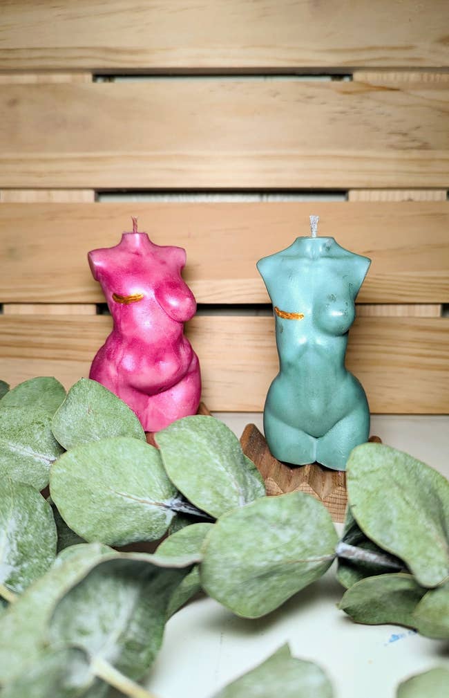 Two torso-shaped candles on a wooden surface with green leaves nearby
