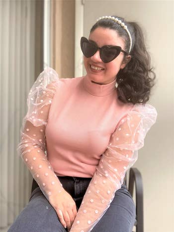Woman in a pink top with sheer polka-dot sleeves and heart-shaped sunglasses, seated, smiling