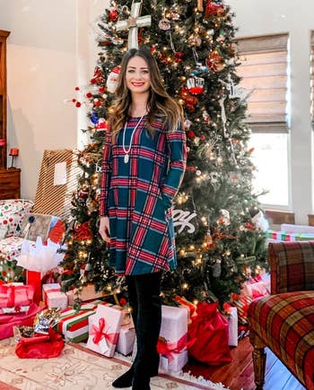 reviewer wearing the green and red plaid dress with black tights and boots in front of a christmas tree