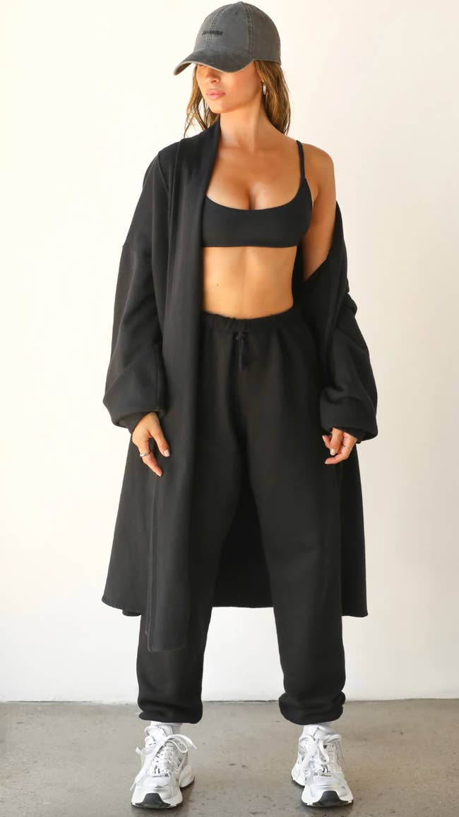 Woman in athleisure: black sports bra, high-waisted sweatpants, long black cardigan, grey cap, and white sneakers