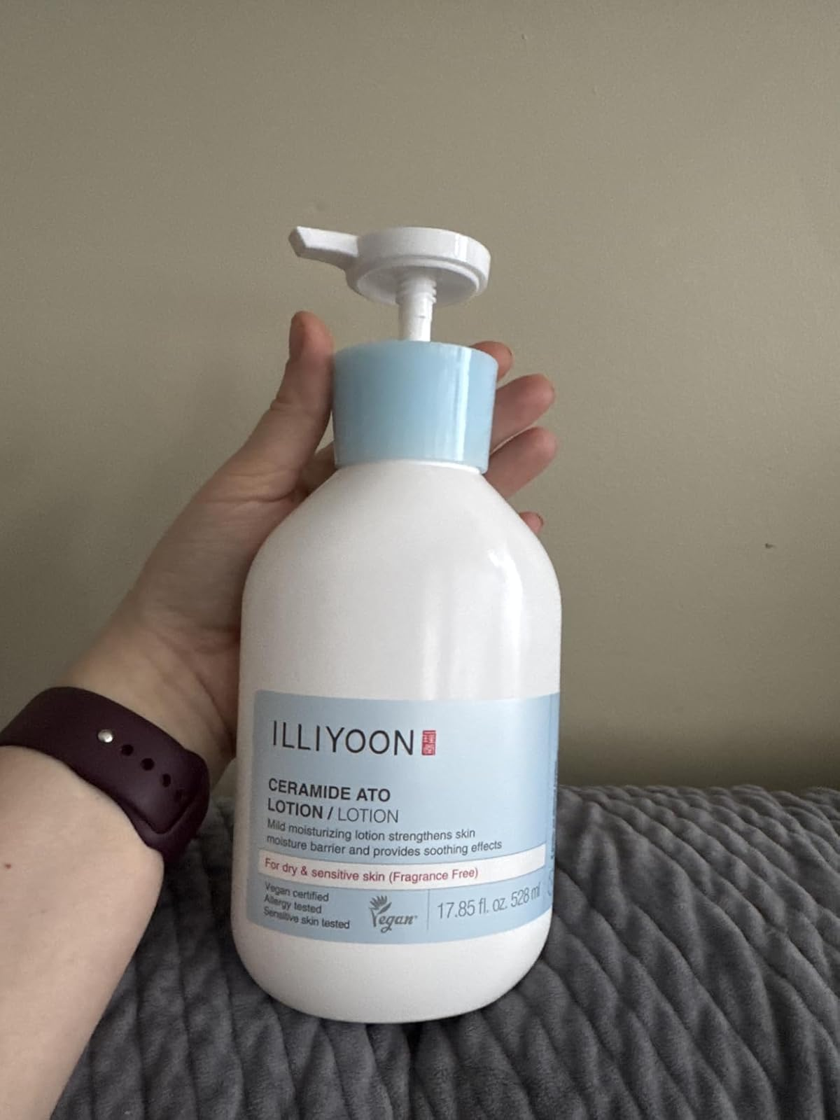 Person holding a vessel  of Illiyoon Ceramide Ato Lotion, labeled for adust  and delicate  skin, connected  a textured surface