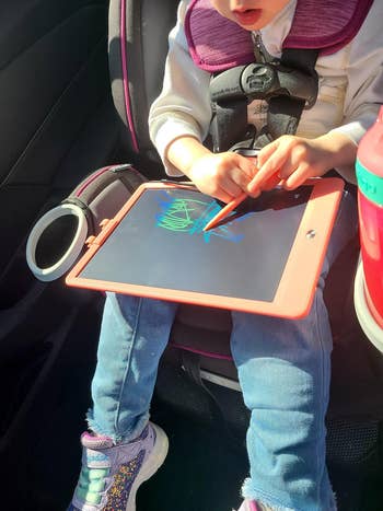 Child drawing on tablet in car seat, using stylus. Jeans and sneakers visible