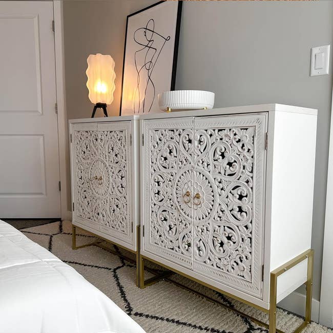 Ornate white cabinets with intricate designs, topped with a lit lamp, a round bowl, and abstract artwork, next to a white door and a bed