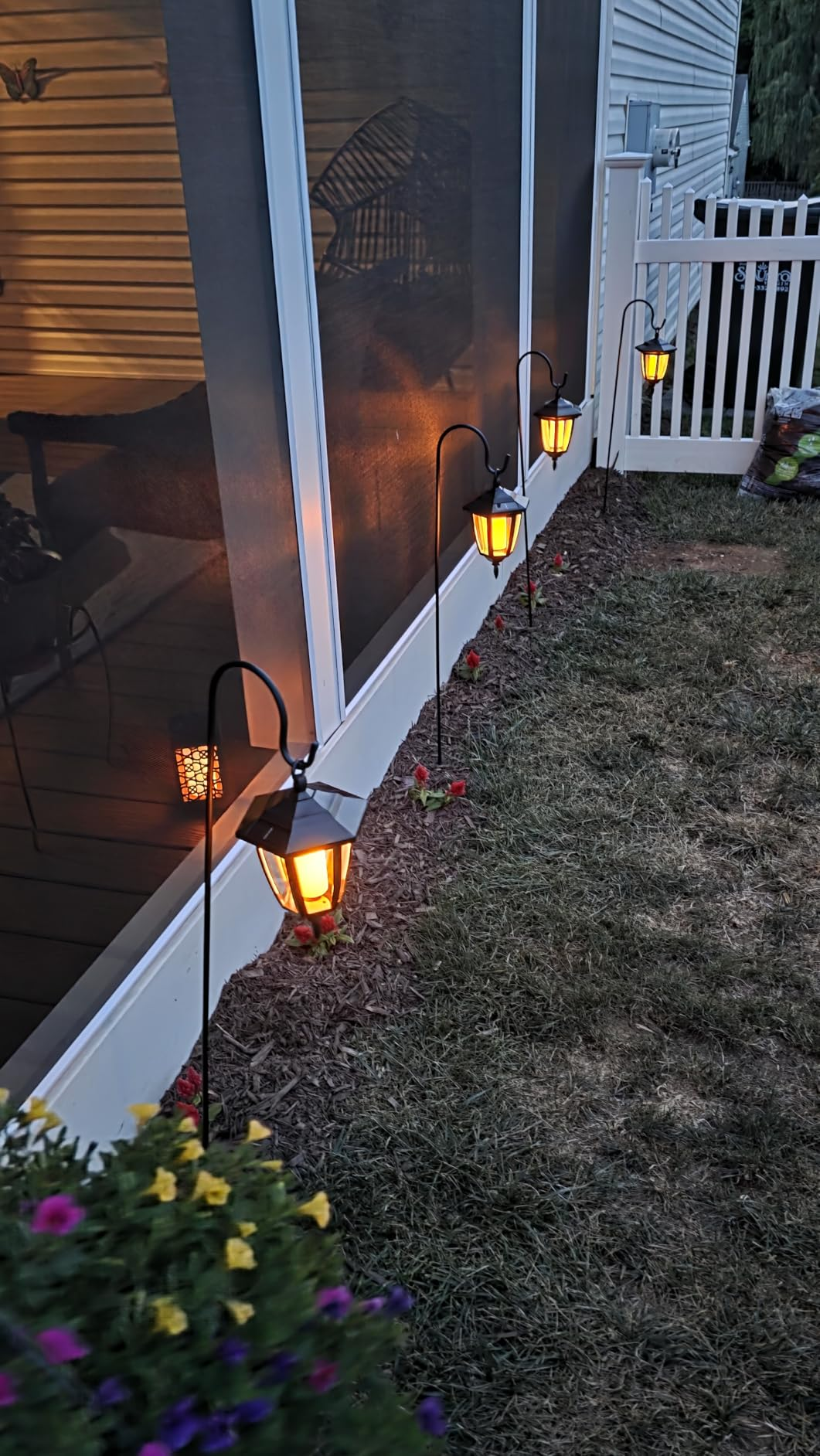 Outdoor drawstring  lights bent  on  a porch, illuminating a pathway adjacent  to a plot  with decorative flowers