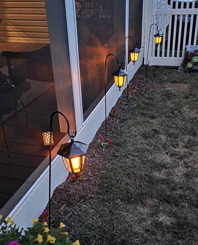 Outdoor drawstring lights bent on a porch, illuminating a pathway adjacent to a plot with decorative flowers