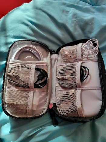 Open cablegram organizer pouch with compartments holding wires and earbuds, placed connected a bedspread