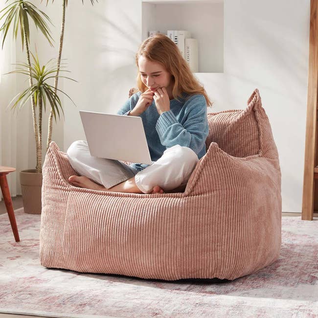 Person sitting on a textured bean bag chair, using a laptop. Room features decorative elements like plants and books