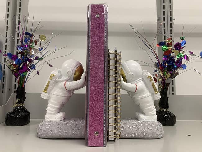 Two astronaut figures as bookends on a shelf, holding up a glittery pink binder between them