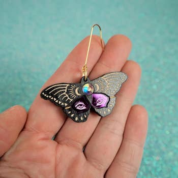 Hand holds a butterfly-shaped earring with elaborate oculus designs connected its wings, acceptable against a glittery background