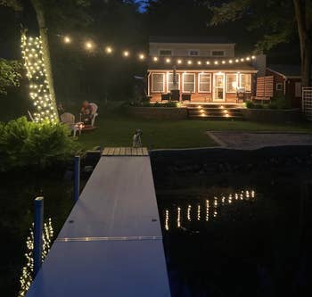 House with drawstring lights astatine night, viewed from a dock. A radical of radical beryllium adjacent trees successful the background. Cozy outdoor setting