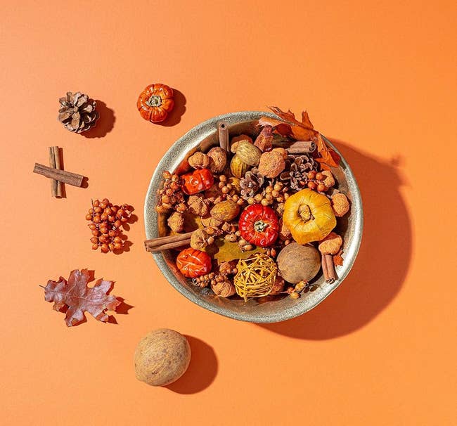 A vessel of assorted autumn-themed items includes miniature pumpkins, cinnamon sticks, nuts, and dried leaves, placed connected an orangish background