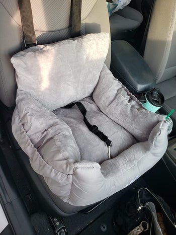 Reviewer image of gray plush dog car seat inside car with two thick straps attached to headrest and a harness clip inside of it