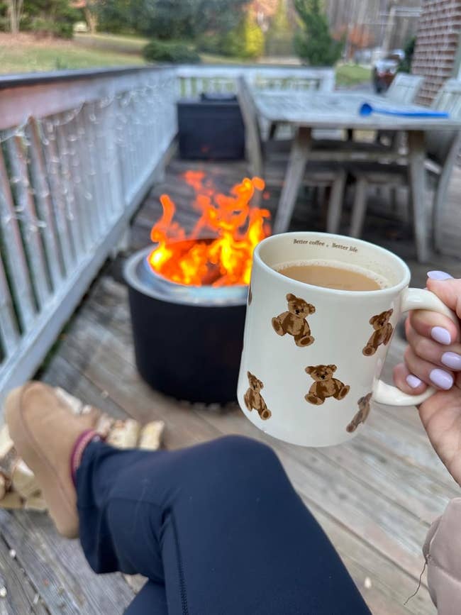 Person relaxing on a deck with a mug depicting teddy bears, near a fire in a metal pit. Wooden chairs and table visible in the background