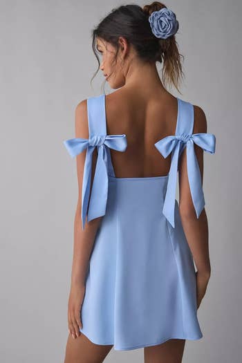 A model in a backless dress with large ribbon bows stands with her back to the camera, featuring a matching hair accessory